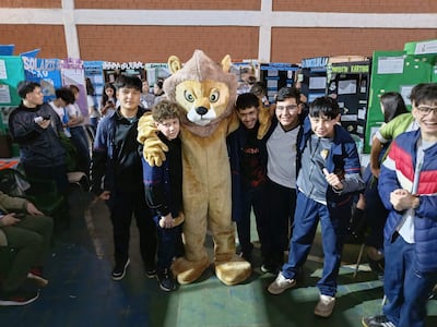 Grupo de seis jóvenes sonrientes en uniformes escolares, rodeando un disfraz de león en un ambiente escolar.