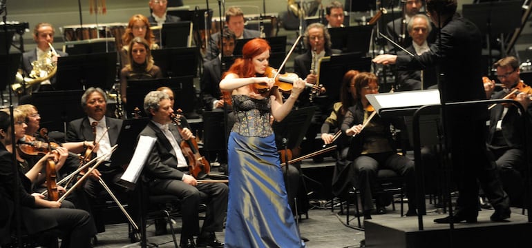 Rachel Kolly, con vestido azul, toca el violín en el centro del escenario, mientras músicos la acompañan.