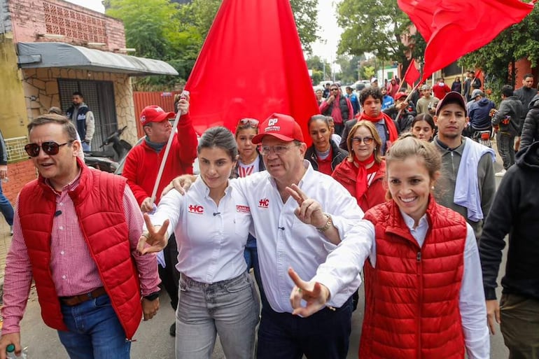 Costanza Troche, sonriente con camisa blanca y logo 'HC', y Camilo Pérez con gorra roja, marchan entre multitudes con banderas rojas.