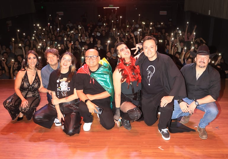 Seis artistas sonríen en el escenario, con vestimentas coloridas, mientras el público ilumina el fondo con sus teléfonos móviles.