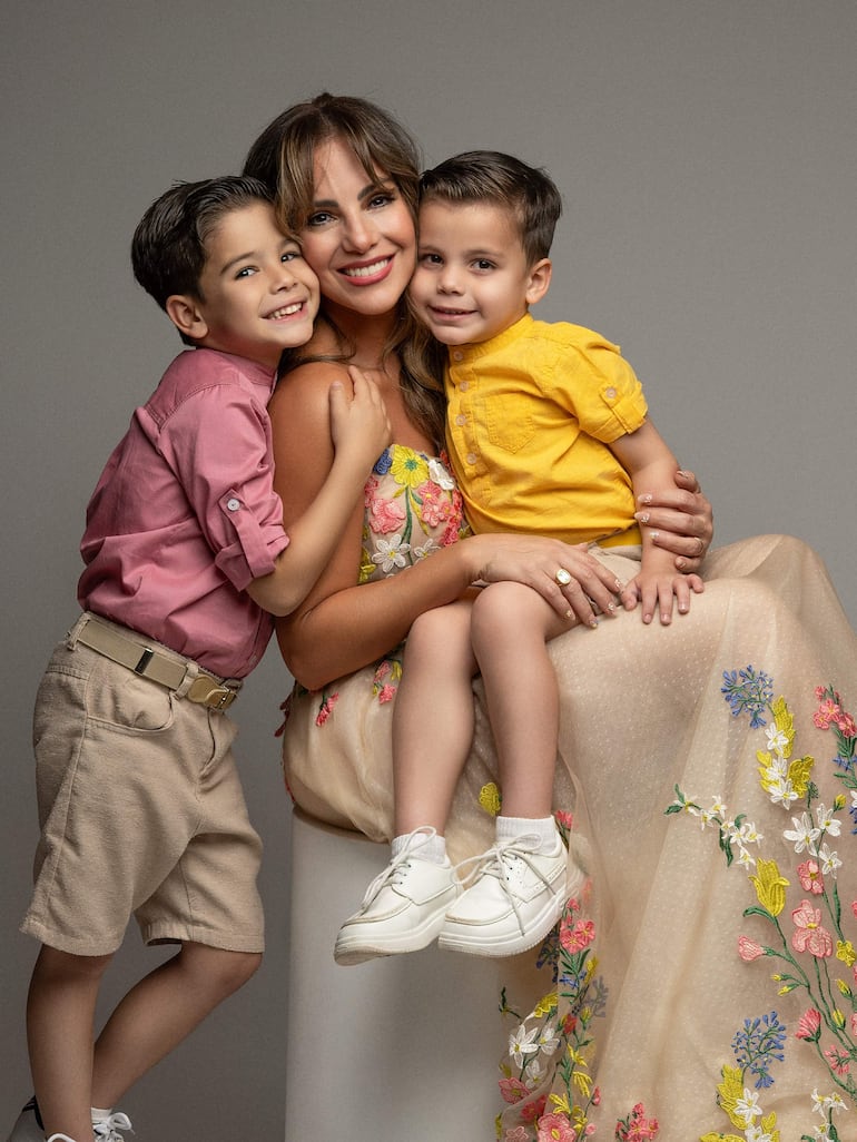 Mujer con vestido claro y bordados rodeada de dos niños, uno con camisa rosa y otro con camiseta amarilla, todos sonrientes.