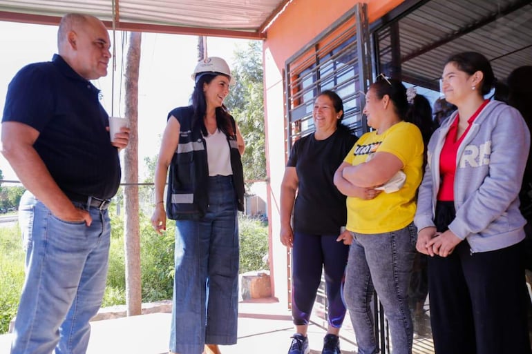 Basilio con camiseta negra y vaso, Claudia con casco y chaqueta de seguridad, sonríen en un entorno soleado y comunitario.