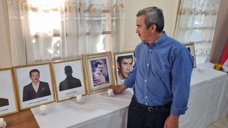 Hombre de pie con camisa azul con puntos blancos observa retratos enmarcados y velas, en ambiente de conmemoración.