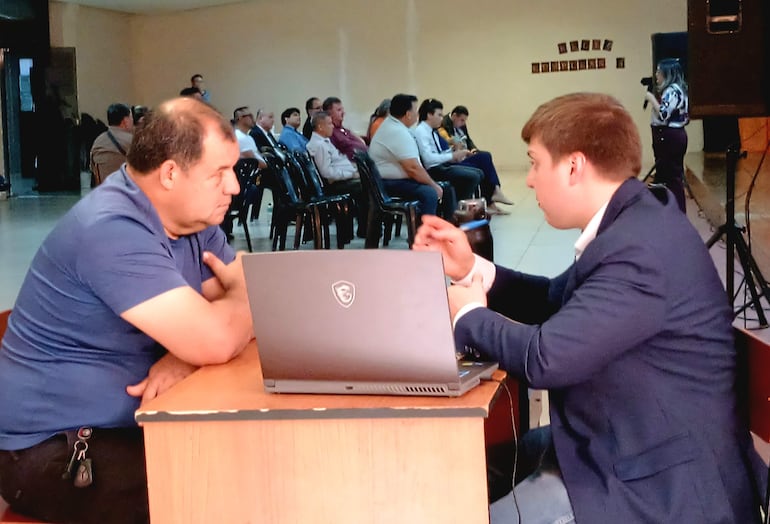 Dos hombres sentados en mesa; uno en camiseta azul escucha al otro con saco. Fondo con público atento.