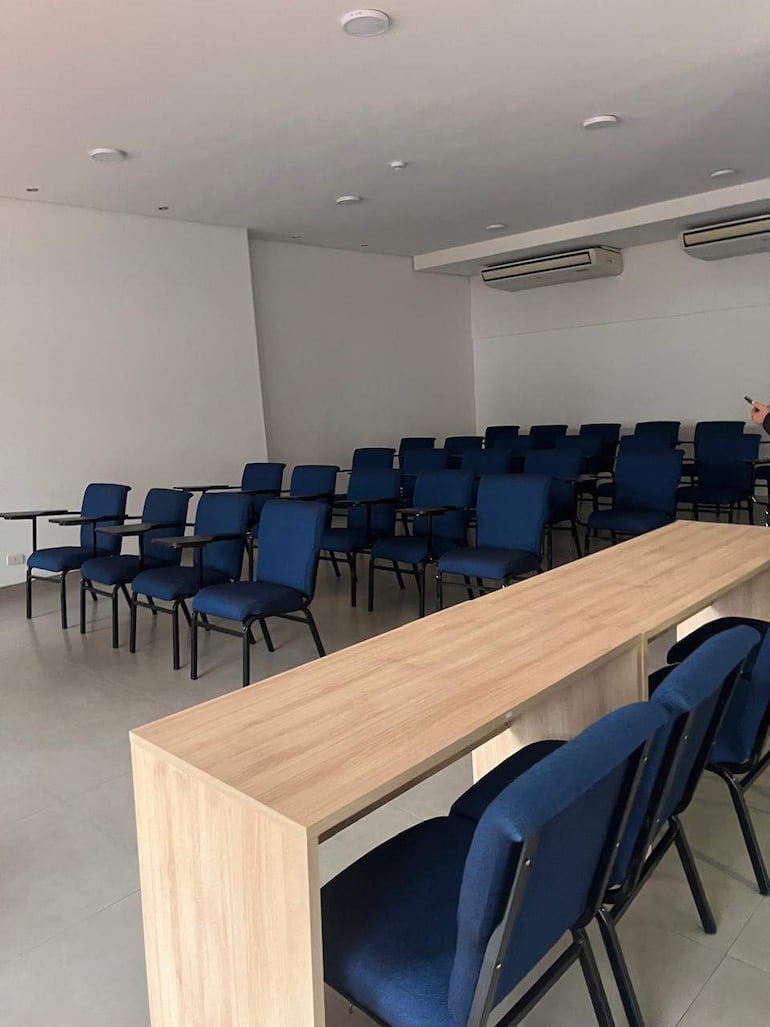 Sala vacía con sillas azules dispuestas en filas y un mostrador de madera. Ambiente moderno y minimalista.