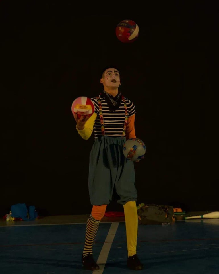 Payaso en vestuario colorido y cara pintada, malabareando pelotas en un ambiente de circo oscuro.