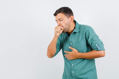 Hombre de pie con camisa verde, tosiendo y cubriendo su boca, en un fondo blanco y sin otros elementos visibles.
