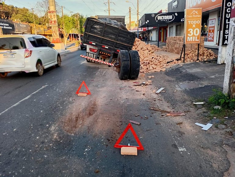 Camión volcado con ladrillos esparcidos en la carretera. Triángulos de advertencia indican peligro en entorno urbano.
