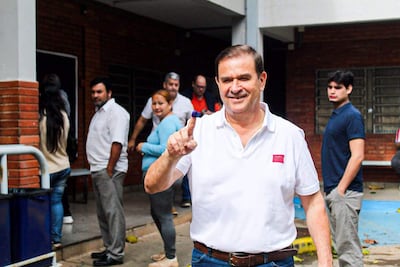 Hombre sonriente con camisa blanca levanta su dedo entintado, rodeado de otros votantes en un ambiente exterior.