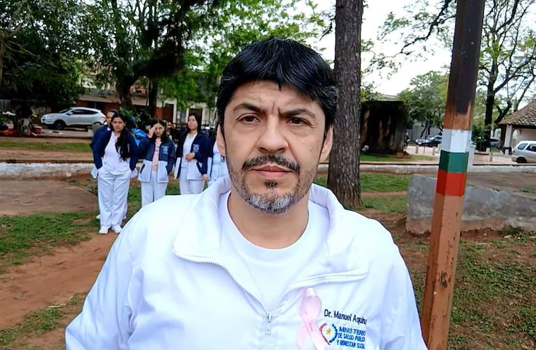 Manuel Aquino con chaqueta blanca de médico, rodeado de mujeres con chaquetas oscuras, en un ambiente verde y natural.