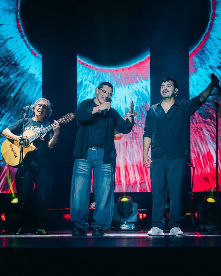 Tres hombres en el escenario: uno toca guitarra, otro canta con gafas y el tercero levanta la mano, todos vestidos de negro.