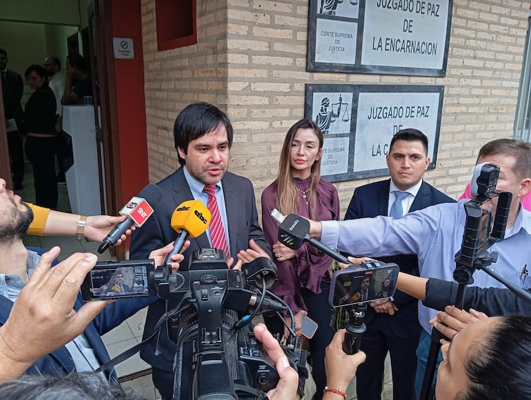 Hombre con traje oscuro y corbata roja sostiene un micrófono, mientras otros escuchan en el Juzgado de Paz de La Encarnación.