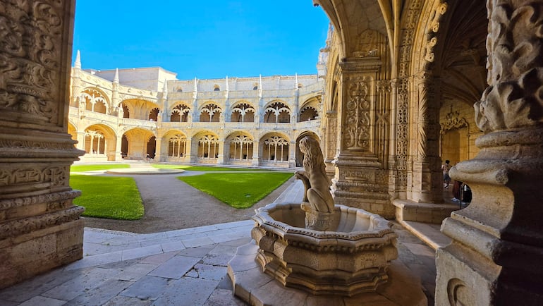 Claustro con arcos góticos, fuente en forma de león y césped verde vibrante bajo un cielo azul.