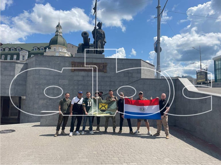 Ocho personas sonrientes, algunas en camisetas militares, sostienen banderas paraguayas frente a un monumento bajo un cielo soleado.
