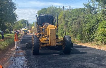 Tres trabajadores, uno con camiseta naranja, operan al lado de una motoniveladora amarilla en una carretera rodeada de vegetación.