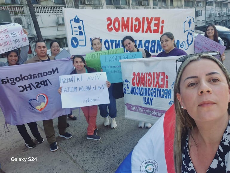 Grupo de diez personas en manifestación, mujer sonriente con blusa negra y blanca sostiene bandera de Paraguay junto a pancartas de diversas demandas.