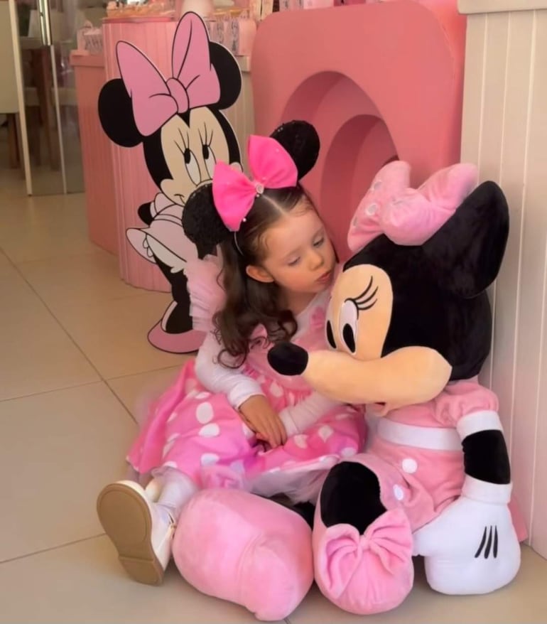 Niña pequeña de vestido rosa y diadema de orejas de ratón, sentada en el suelo viendo un muñeco de Minnie Mouse.