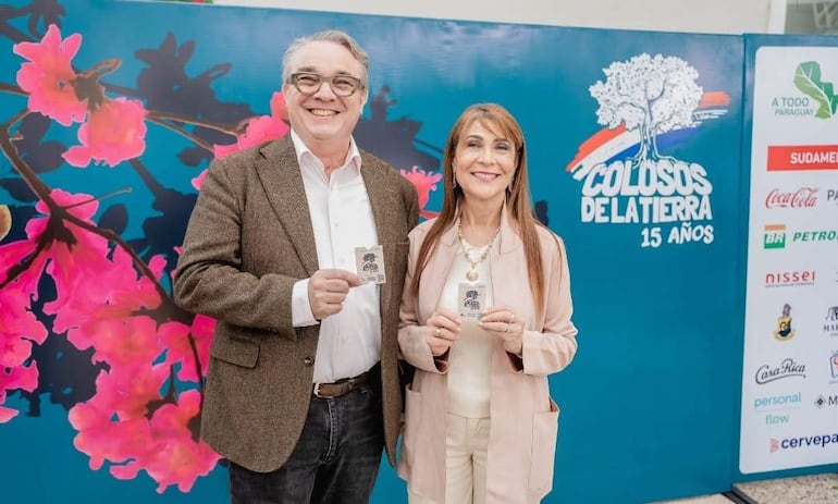 Hombre canoso con blazer marrón y mujer con abrigo claro sonríen frente a fondo floral, sosteniendo tarjetas del evento.
