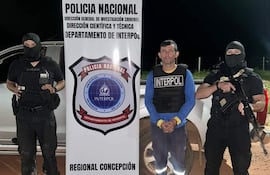 Hombre con camiseta azul y chaleco de Interpol, custodiado por dos hombres armados en un entorno rural con vehículo policial de fondo.