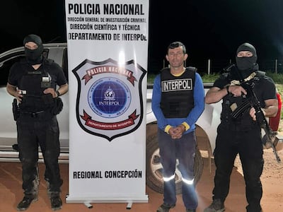 Hombre con camiseta azul y chaleco de Interpol, custodiado por dos hombres armados en un entorno rural con vehículo policial de fondo.