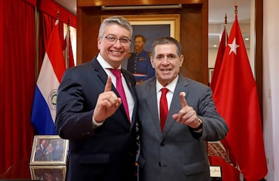 Horacio Cartes en traje negro y corbata roja, sonriente, junto a Ariel Ojeda en traje gris, ambos levantando un dedo en un ambiente formal.