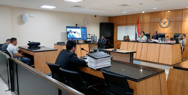 Sala de audiencias con Barrios en camiseta blanca y abogado en traje oscuro, mientras un monitor muestra la audiencia virtual.