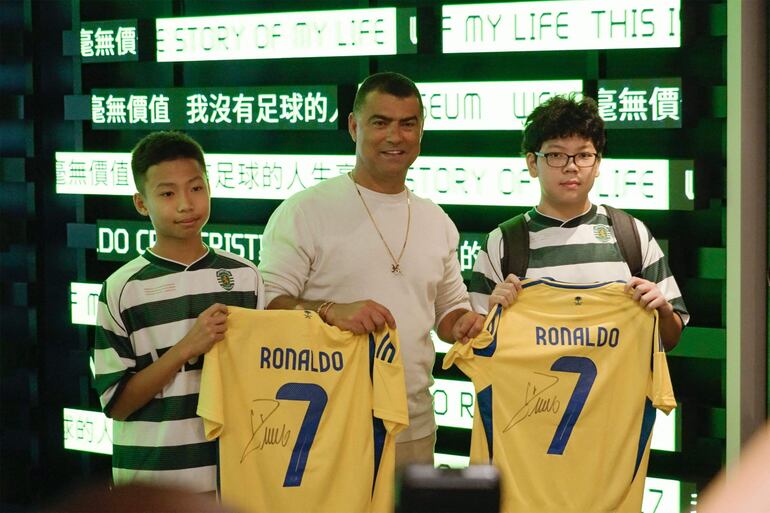 Esta captura de pantalla de un vídeo de AFPTV muestra a dos aficionados recibiendo camisetas autografiadas de Cristiano Ronaldo mientras posan con el hermano del futbolista, Hugo dos Santos Aveiro (centro), durante la inauguración del Museo CR7 LIFE en Hong Kong.