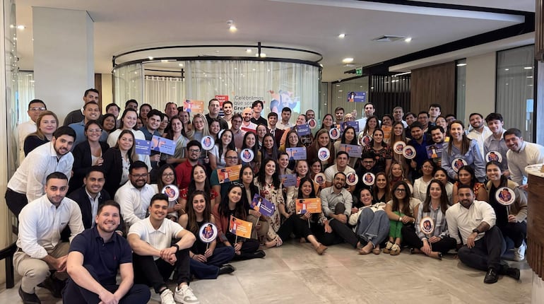 Diverse grupo de 80 personas sonríe y sostiene carteles en un ambiente festivo, con detalles coloridos en la ropa.