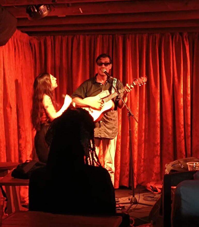 Hombre con gafas oscuras toca guitarra al micrófono, mientras mujer de cabello largo canta en ambiente cálido con cortinas rojas.