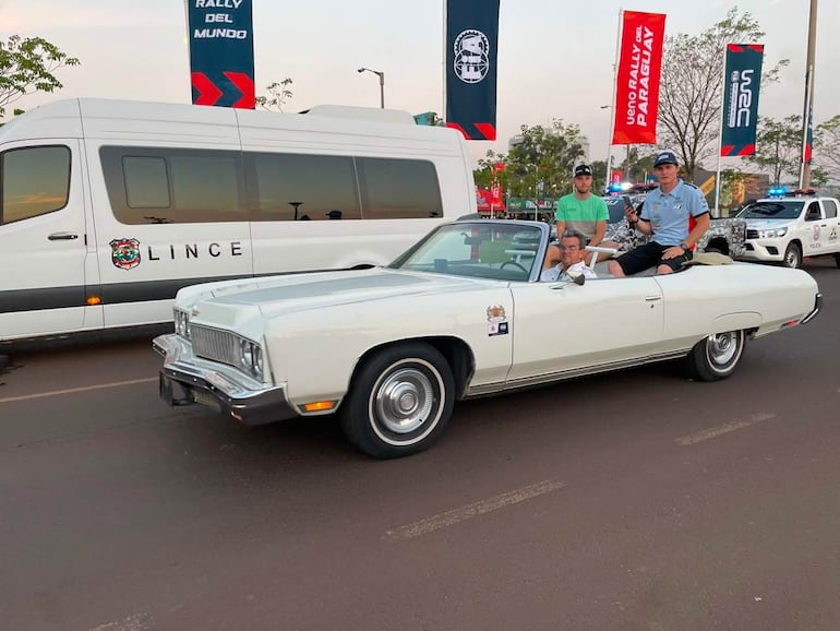 Hombres en un convertible blanco durante un desfile, sonrientes y emocionados, con furgoneta 'LINCE' y banderas del rally de fondo.