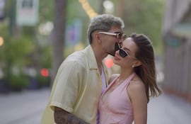 Julio Enciso con camiseta amarilla besa la frente de Melissa Cardona, que lleva un vestido rosa y gafas de sol, en un ambiente urbano.