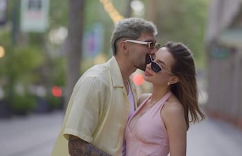 Julio Enciso con camiseta amarilla besa la frente de Melissa Cardona, que lleva un vestido rosa y gafas de sol, en un ambiente urbano.
