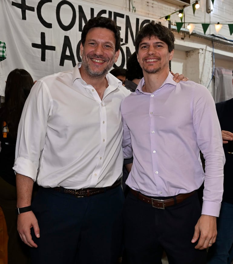 Hombres sonrientes, uno en camisa blanca y otro en camisa púrpura, abrazados en un entorno colorido con pancarta sobre conciencia.