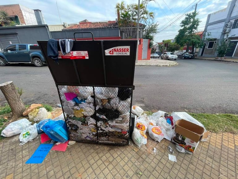 Contenedor negro desbordado de basura en esquina de calle, rodeado de bolsas y cartones, en área residencial de Asunción.