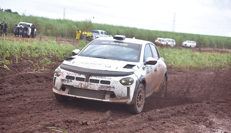 En la imagen se observa un vehículo todoterreno, claramente utilizado en una competición de rally. El auto, parcialmente cubierto de barro, tiene el número 89 visible en su lateral. Se encuentra en un terreno fangoso, característico de un circuito de rally, rodeado por vegetación de caña de azúcar.
No hay personas visibles en la imagen que sean reconocibles como figuras públicas. El ambiente es exterior y parece ser un circuito rural donde se lleva a cabo una carrera.
Al fondo, se pueden distinguir vehículos estacionados, junto con personas en el área de espectadores, aunque no se pueden identificar claramente sus características.
No hay elementos visuales que indiquen la presencia de un equipo de fútbol, ni lugares icónicos o estadios en la imagen. La acción principal es el vehículo competidor moviéndose por el terreno barroso.