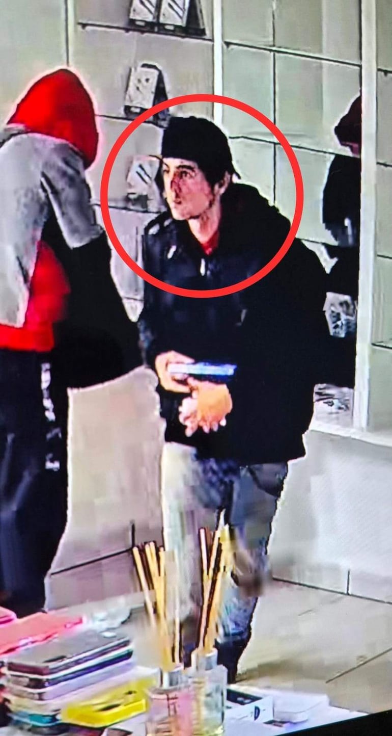 Joven con chaqueta negra y expresión neutral conversando con otra persona en un entorno de tienda, con estantes de objetos detrás.