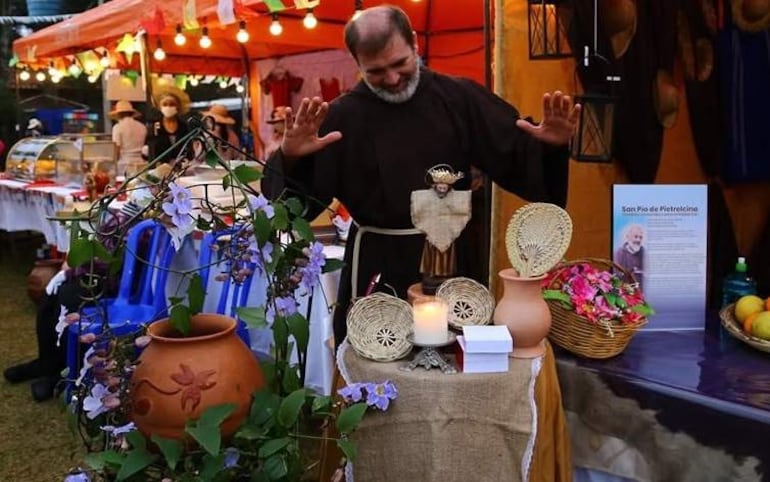 Fraile con hábito oscuro levanta las manos en bendición frente a un altar decorado, rodeado de mesas con comida y luces festivas.