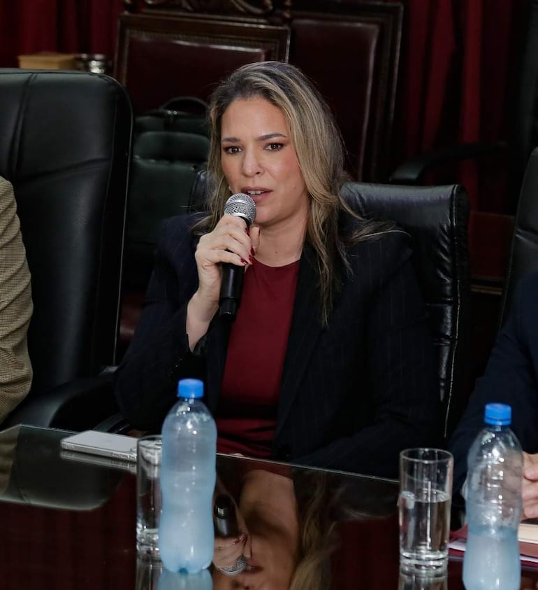 Cecilia Rodríguez, con blazer oscura y blusa roja, habla sosteniendo un micrófono mientras otros hombres la observan en un salón formal.