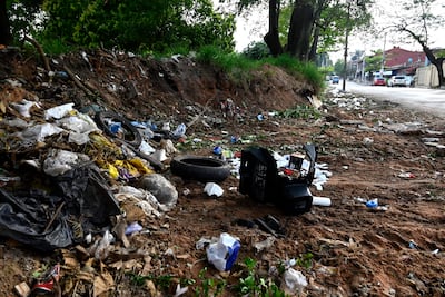 Área desolada con acumulación de basura, incluyendo neumático y mueble, rodeada de vegetación y edificaciones.