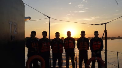 Seis hombres alineados en un barco, visten chalecos salvavidas naranjas, con una puesta de sol de fondo.