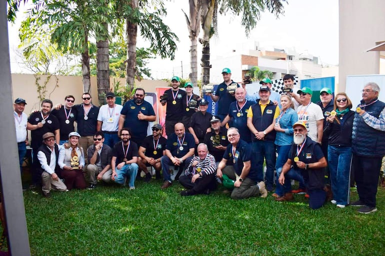Grupo de personas con medallas y trofeo dorado, algunos de pie y otros agachados, en ambiente festivo con plantas alrededor.