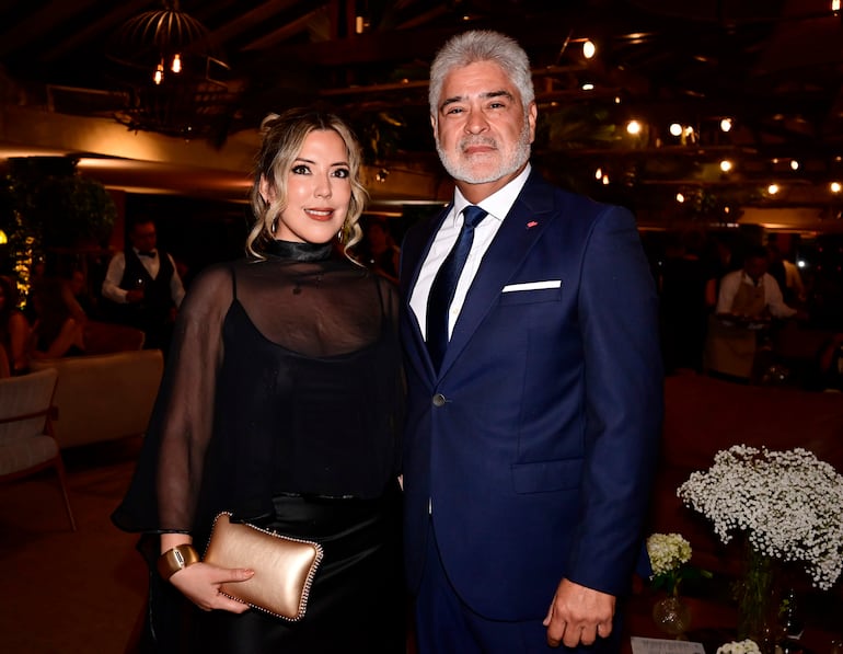 Mujer con vestido negro y bolso dorado junto a hombre canoso en traje azul, ambos sonriendo en un ambiente elegante.
