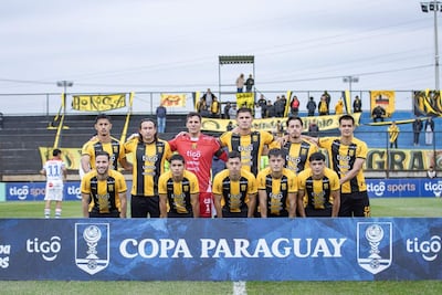 Once jugadores del Club Guaraní lucen camisetas amarillas y negras, sonriendo en el estadio con banners de apoyo en el fondo.