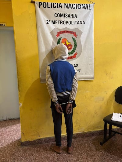 Hombre de espaldas con capucha y grilletes, en comisaría amarilla con documentos sobre mesa y silla vacía.