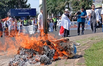 Manifestantes con batas blancas sostienen una bandera paraguaya, con fuego en la acera y policías controlando el orden.