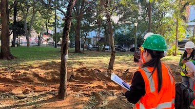 Cinco trabajadores en la Plaza Uruguaya, vestidos con chalecos de seguridad naranjas y cascos, uno sostiene un móvil mientras toma notas.