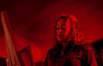 Mujer de cabello rizado y suelto, con chaqueta oscura, frente a un caballete en un ambiente iluminado con luces rojas.