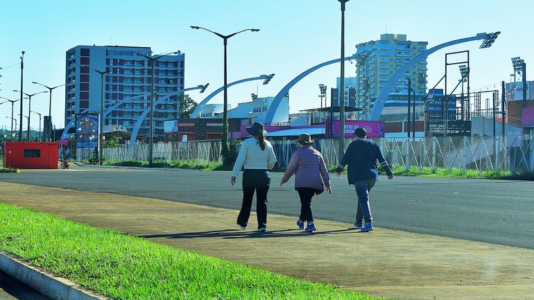 Tres personas caminando en la costanera: mujer con abrigo rosa, hombre con abrigo oscuro y mujer con chaqueta clara en un día soleado.