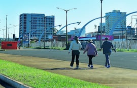 Tres personas caminando en la costanera: mujer con abrigo rosa, hombre con abrigo oscuro y mujer con chaqueta clara en un día soleado.