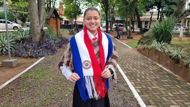 Mujer sonriente con poncho rojo, blanco y azul en un entorno con árboles y vereda pavimentada.